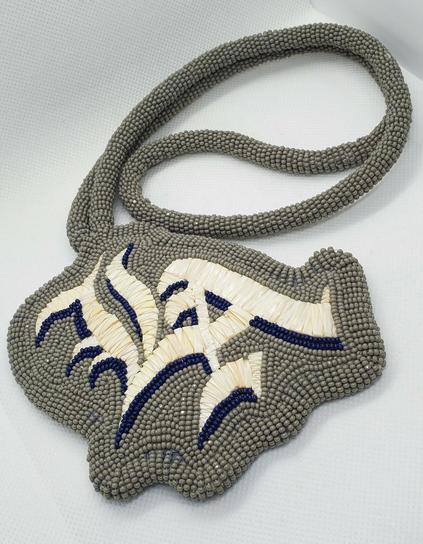 the same beaded gray medallion made by either kiamichi or her daughter with the letters LA in blue and white in the center in old english font made from porcupine quills. this time it's on a white background so the viewer can see the craftsmanship more clearly