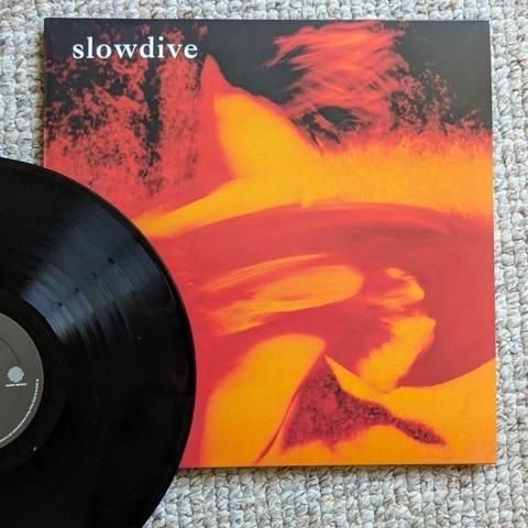 Eine schwarze Vinyl-Schallplatte neben ihrem Albumcover mit abstraktem orangefarbenem und rotem Artwork und dem Text "slowdive" am oberen Rand. Der Hintergrund ist strukturiert und in hellen Farben gehalten.