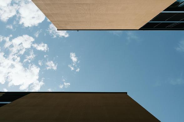 Upward view between two modern buildings with smooth, tan-colored walls and sharp edges, framing a patch of blue sky scattered with white clouds. The clean lines and contrast create a minimalist architectural composition.

