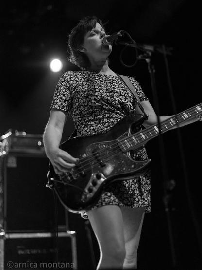 A musician performs on stage, holding a bass guitar and singing into a microphone. She wears a floral-patterned dress, and the image is in black and white, capturing a live music atmosphere with stage lighting in the background.