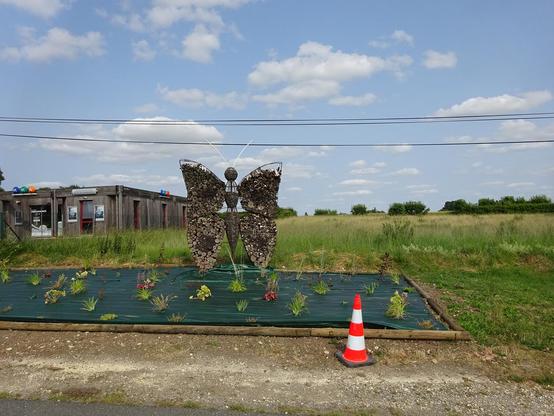 un papillon géant qui est une cabane à insectes sur un parterre bâché avec quelques plantation et un cône de chantier devant