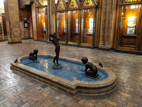 A bronze fountain sculpture in the lobby of the 1927 Fine Arts Building in Los Angeles, California, featuring three figures by sculptor Burt Johnson. Two children are positioned at the base playing in a shallow blue-tiled pool, while a central figure stands gracefully playing a flute, with ornate wooden display cases and intricate architectural details in the background. The pool enclosure, floor and columns are tiles by by Pasadena tilemaker Ernest A. Batchelder.