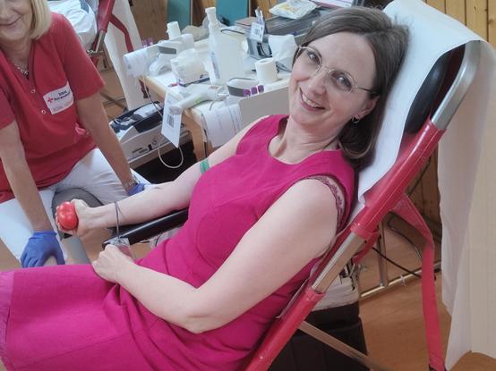 Dr. Bialk entspannt auf einem Stuhl beim Blutspenden. Im rechten Arm liegt der Zugang, im Hintergrund befindet sich ein Tischchen mit medizinischem Instrumentarium. Links im Bild hinter dem Stuhl die durchführende DRK-Schwester mit freundlichem Lächeln. Sie trägt ein rotes T-Shirt und weiße Hosen.