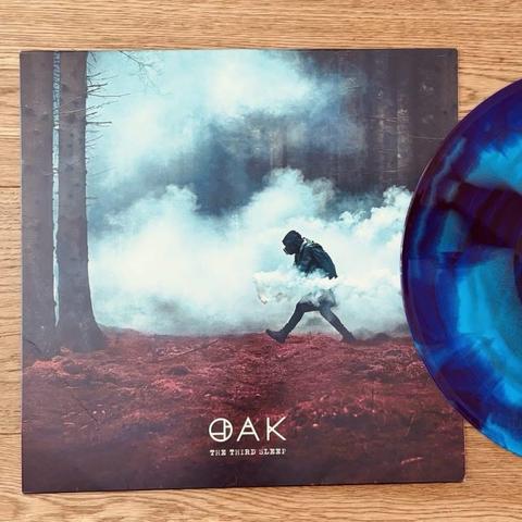 Das Vinyl-Schallplattencover zeigt eine neblige Waldszene mit einer Person, die durch den Nebel geht. Das Artwork besteht aus dunklen Bäumen und einem rötlichen Grund, auf dem der Titel "OAK - THE THIRD SLEEP" prangt. Ein blau und schwarz marmoriertes Vinyl ist rechts daneben zu sehen.