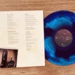 Eine blau und schwarz marmorierte Schallplatte liegt neben einem Album, auf dem Songtexte aufgedruckt sind. Der Hintergrund ist eine Holzfläche.