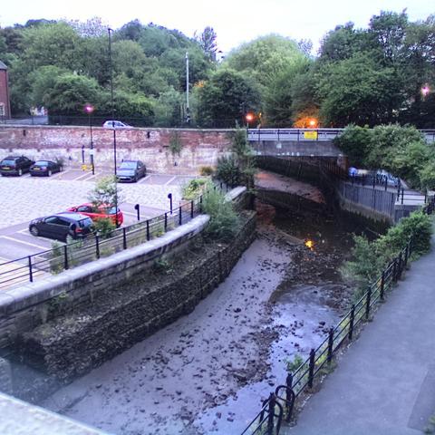 Photograph of the tidal stretch of Ouseburn River at south side of Ouseburn Bridge in Newcastle upon Tyne. To the left (south-west) of the bridge is Cut Bank, and to the right (north-east) is Byker Bank. In the foreground is the pedestrian Riverside Walk and the photo also spans some surrounding built-up areas and trees, with sky in the background. Still image captured from webcam at 21:48 hrs on Tuesday 24 June 2025.