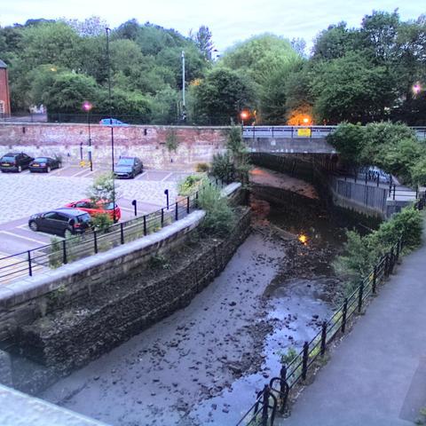 Photograph of the tidal stretch of Ouseburn River at south side of Ouseburn Bridge in Newcastle upon Tyne. To the left (south-west) of the bridge is Cut Bank, and to the right (north-east) is Byker Bank. In the foreground is the pedestrian Riverside Walk and the photo also spans some surrounding built-up areas and trees, with sky in the background. Still image captured from webcam at 21:52 hrs on Tuesday 24 June 2025.