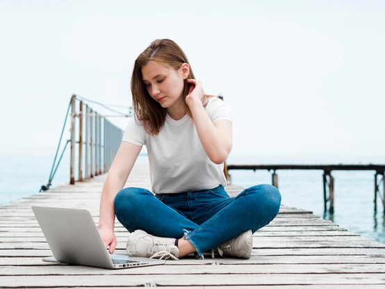 a woman, sitting on a pier, with her right hand on the keyboard of a laptop computer that is sitting next to her on the pier