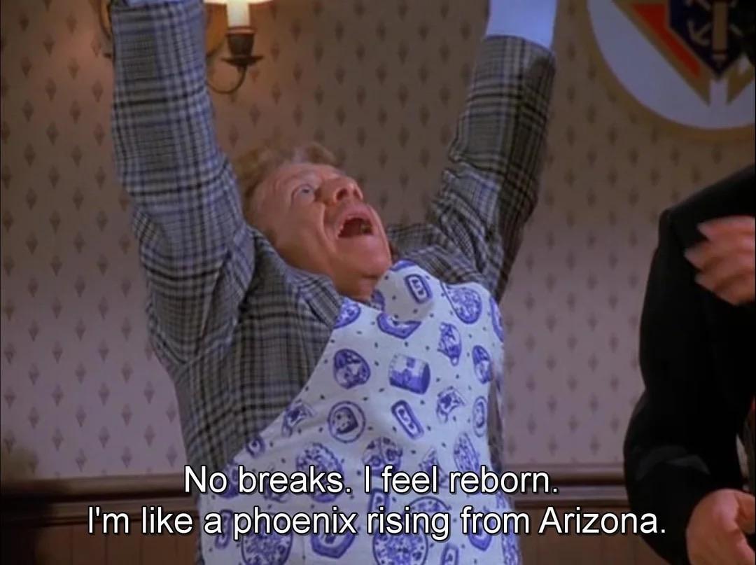Frank Costanza excitedly raises his arms, expressing joy and a sense of renewal. He wears a blue patterned shirt beneath a gray plaid jacket. The scene includes a wooden interior with a lamp and wallpaper.  "No breaks. I feel reborn. I'm like a phoenix rising from Arizona."