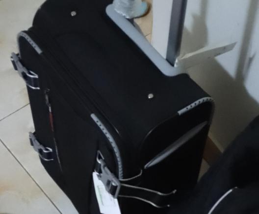 The image shows a black suitcase with a hard shell and a soft fabric exterior. The suitcase is standing upright on a tiled floor, with a white wall in the background. It features a gray handle on top and a side zipper pocket with a red accent. There are two gray buckles on the side, likely for securing the contents. A luggage tag is attached to the side, with a white background and green text. The suitcase has a silver trim along the edges and a gray accent on the top. A plastic-wrapped item is partially visible on top of the suitcase. The lighting is dim, and the overall image is slightly blurry.

Provided by @altbot, generated privately and locally using Ovis2-8B