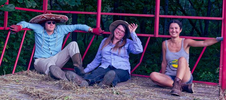 Three people sit in a red metal hay wagon with dark forest in the background and a wood floor littered with loose grass.  Left to right:  (1) man wearing a blue shirt, cream pants, red gloves, dark glasses and decorated red sombrero, (2) woman with blue shirt and pants, long hair and hat and (3) younger woman with shorts, tank top, and tied up hair