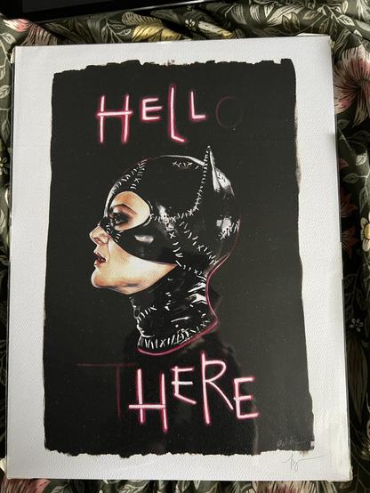 Picture showing a print, a roughly rectangular black portrait shaped background on which are words in neon at top and bottom which when lit, would spell out “HELLO THERE”
In the middle of the words is a profile art of Michelle Pfeiffer as “Catwoman” from “Batman Returns” (a light skinned femme presenting woman wearing a jaggedly sewn together rubber/latex hood with cat ears)
Per the movie scene, the neon letters have been damaged and the caption now reads:

“HELL HERE”