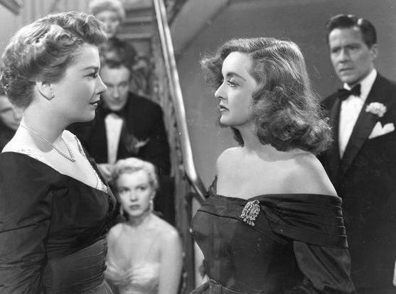 Schwarzweiss Filmstil aus dem Film "All about Eve". Im Vordergrund schauen sich zwei Frauen an im Gespräch. Im Hintergrund sitzen Menschen auf einer Treppe. Am rechten Bildrand steht ein Herr im Anzug mit Fliege.