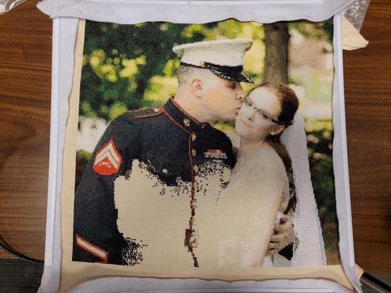 Marine with a short haircut on the left with a white hat and black or very dark blue uniform. Om the left side on his arm theres a red patch with two light yellow chevrons and a pair of rifles crossed below it.  Bride in a white dress on cream color fabric with a white and silver veil.  There are greens and Browns making up the wooded background with a tan colored building behind some trees on the right and some darker foliage in the top left.