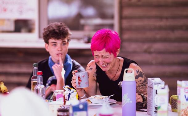 A woman with pink hair sits on a bench, laughing. Left besisde her a young boy with short brown hair