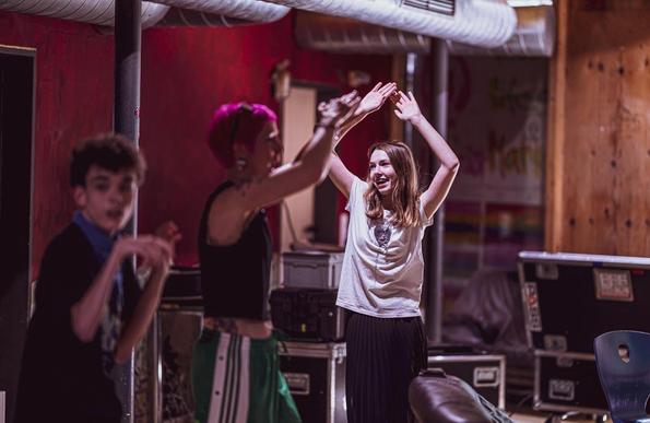 A woman with pink hair smiles at a girl with long brown hair, the are dancing. On the left looks a boy in the camaera, he is dancing too
