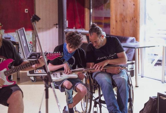 A man in a wheelchair plays bass guitar alongside two other men, creating music together in a lively setting.