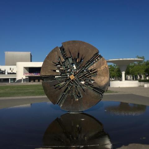 Large abstract disc-shaped bronze sculpture in the bright summer sun. It is reflected in the shallow water it stands in, with the white marble entrance building of the Staatstheater Darmstadt visible behind it. The sky above is deep blue and cloudless. «Grande Disco» by Arnaldo Pomodoro (23 June 1926 – 22 June 2025) on Georg-Büchner-Platz on June 25, 2025.
Photo by Stefan Romero Grieser @operaimmagine, CC BY-SA 4.0.