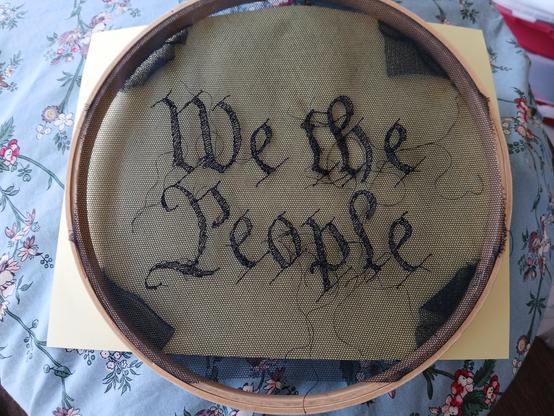 An embroidery hoop holds a piece of black netting. The hoop is about a 10 inch diameter. You can fit your face in it, for example.

Embroidered on the center of the veil is the phrase "We the People" copied from the original script of the US Constitution. It looks a little scratchy like the original print copies because of the way the embroidery went. The letters are outlined in a slightly heavier thread. 