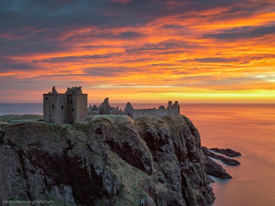 A photograph of Dunnottar Castle in Aberdeenshire, Scotland, perched on a rugged cliff overlooking the sea. The sky behind it blazes with fiery shades of orange, red, and gold from the rising sun. The castle itself appears mostly in shadow, its crumbling stone walls and towers forming a dark silhouette against the vivid dawn. The dramatic contrast between the glowing sky and the stark outline of the ruins highlights the wild beauty of the coastline and the imposing presence of the ancient fortress.