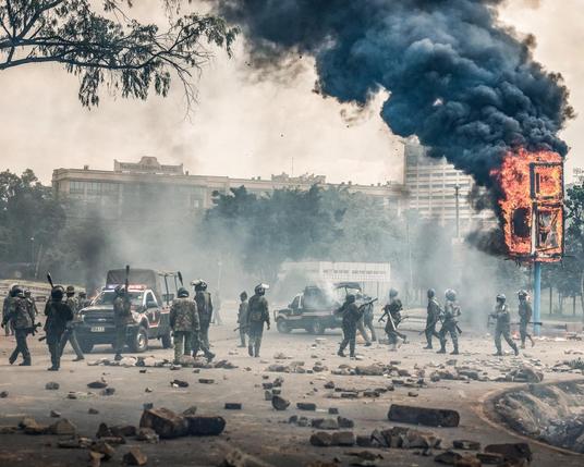 The image depicts a chaotic urban scene with a large plume of black smoke rising from a burning structure on the right side. The structure appears to be a billboard or a large signpost engulfed in flames. In the foreground, a group of uniformed personnel, likely riot police, are seen walking across the street, some carrying batons and wearing helmets. They are positioned in front of a police vehicle with flashing lights. The street is littered with rocks and debris, indicating recent unrest. In the background, buildings and trees are visible, partially obscured by smoke. The overall atmosphere is tense and suggests a situation of civil unrest or protest.