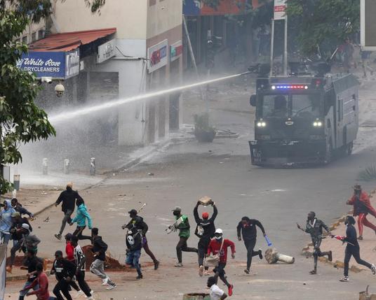 The image depicts a street scene during a protest or riot. In the foreground, a group of people, mostly young adults, are seen running away from a police vehicle equipped with a water cannon. The water cannon is spraying a powerful stream of water across the street. The individuals are dressed in casual clothing, with some wearing masks and others carrying objects like sticks and rocks, suggesting they are prepared for confrontation. The background shows a commercial area with buildings, including a dry cleaner's shop with a sign that reads "White Rose Drycleaners." The atmosphere is tense, with debris scattered on the ground and a sense of urgency among the people. The police vehicle is marked with blue and red lights, indicating it is part of a law enforcement response. The scene is chaotic, with people in various states of motion, some crouching and others running, as they attempt to evade the water spray.