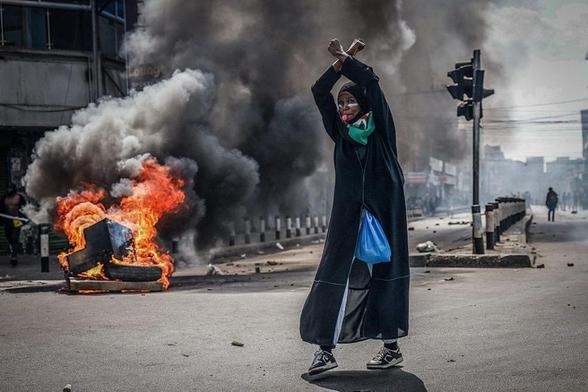 The image depicts a street scene with a person standing in the foreground. The individual is wearing a long black garment with a blue bag hanging from the side, and a Kenyan flag scarf around their neck. They have their arms raised above their head, with fists clenched, suggesting a gesture of protest or defiance. The background shows a street engulfed in thick black smoke, with flames visible from a tire fire, indicating a possible protest or unrest. The street is empty except for the person and a few distant figures, and there are traffic lights and buildings visible in the background. The overall atmosphere is tense and chaotic, with the smoke and fire adding to the sense of disorder.