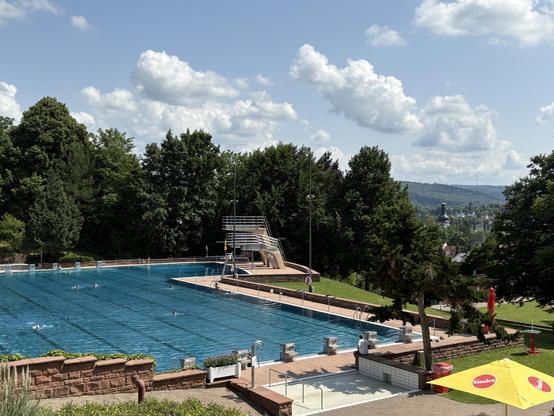 Lahrer Terrassenbad mit klarem blauem Wasser, umgeben von grünen Bäumen. Einige Menschen schwimmen, während andere am Beckenrand sitzen. Im Hintergrund sind ein Sprungturm, Hügel und Wolken am Himmel sichtbar.