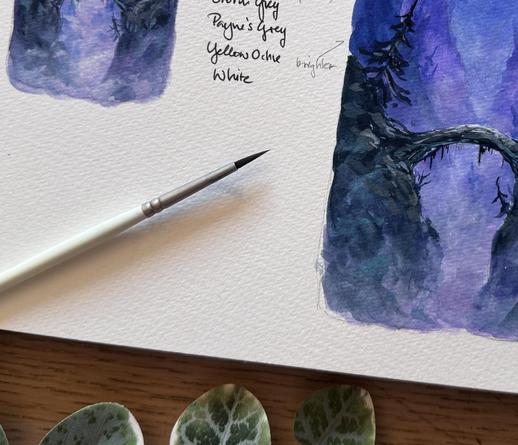 A snippet of a sketchbook, showing bits of two differently sized renditions of a purplish mountain scenery. A paint brush is placed on the sketchbook, and a few small leaves next to it.