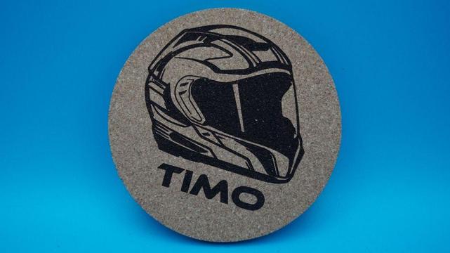 Ein runder Korkuntersetzer mit einem stilisierten Motorradhelm und dem darunter gravierten Namen 'Timo'. Die Gravur ist tiefschwarz und präzise, das Motiv modern und klar. Aufgenommen vor neutralem, blauem Hintergrund.