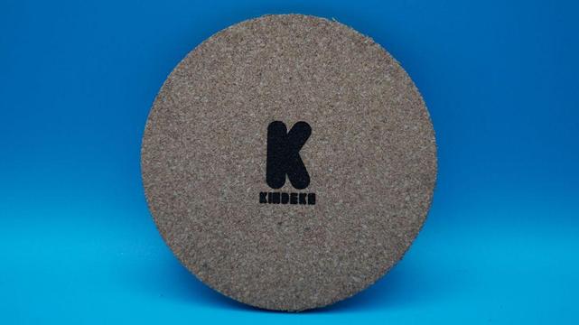 Runder Korkuntersetzer mit zentral graviertem schwarzen Kindeko-Logo. Das Logo besteht aus einem dicken, verspielten 'K' und dem Schriftzug 'KINDEKO' darunter. Die Gravur ist kontrastreich und präzise ausgeführt.