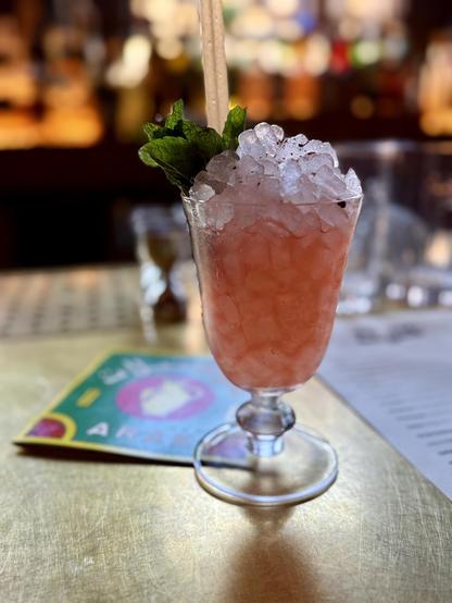 A pink hued cocktail on a brass colored bar with a bunch of mint. The cocktail has arak and tequila and sumac. Very refreshing. 