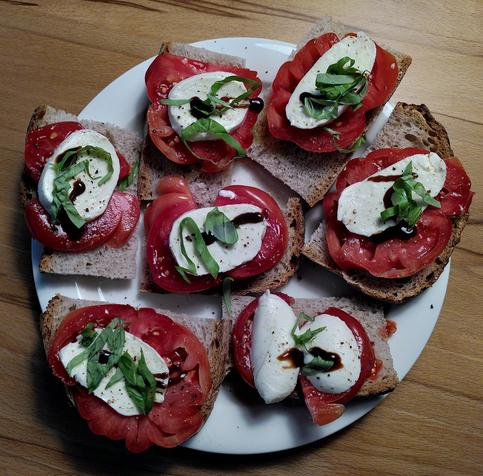 7x Tomate-Mozzarella mit Basilikiumstreifef auf Holzofenbrot liegen auf einem weißen Teller, der auf einer Holzplatte steht.