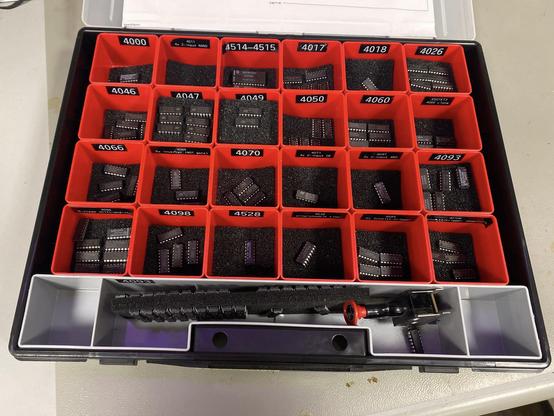 A black plastic carrying case with red square insert compartments. Each compartment has several small integrated circuits - all of the 4000 series - and each compartment (3x18) is carefully labelled with the chip code(s) as white numbers printed on black label tape. 