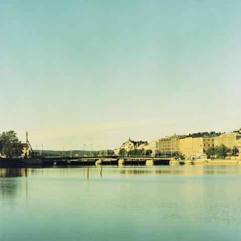 Bridge across a wide canal in a small city on a sunny day. Buldings on both sides. Pastel blue/green colour palette in the sky and water.