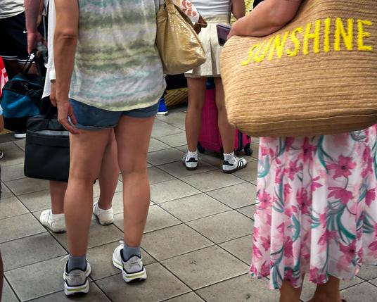 Color photo of a number of people congregated at a subway platform. THey are all facing away from the camera and all are depicted from the shoulders down, no one’s face is showing. A person on the right, wearing a pink and green floral dress has a wicker tote bag slung over their shoulder. On its side is the word, “SUNSHINE” in all capital, yellow stitching. 