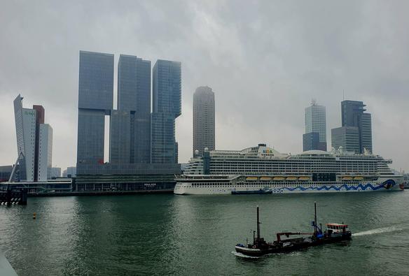Skyline mit modernen Hochhäusern an einem bewölkten Tag. Im Vordergrund befindet sich ein großes Kreuzfahrtschiff mit dem Schriftzug "AIDA" am Heck, das an einem Hafen vor Anker liegt. Neben dem Kreuzfahrtschiff fährt ein kleinerer, dunkler Schlepper mit einem roten Dach über das Wasser. Die Hintergrundlandschaft wird von mehreren Wolkenkratzern dominiert, darunter ein auffälliges Gebäude mit zwei verbundenen Türmen. Die Farben des Bildes sind gedämpft, mit einem dominierenden Grau des Himmels …