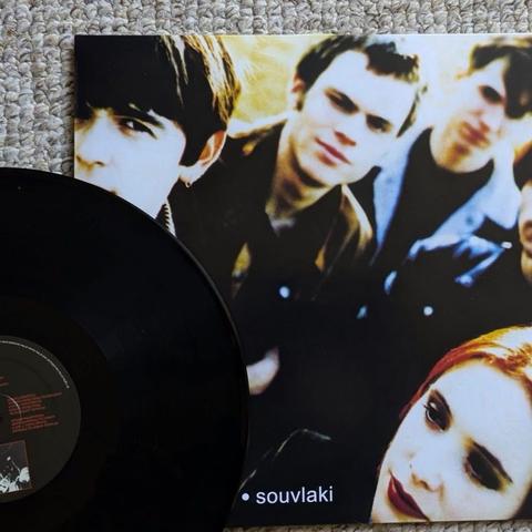 Eine Vinyl-Schallplatte mit dem Cover des Albums "Souvlaki" von der Band Slowdive. Das Cover zeigt eine Gruppe von Menschen vor einem weichen, verschwommenen Hintergrund. Die Schallplatte selbst ist schwarz und liegt neben dem Albumcover.