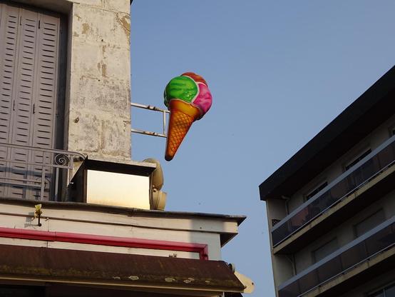 une enseigne représentant une glace trois boules dans un cornet.
Une boule verte, une rose et une orange au dessus.
Sur le côté d'un immeuble à la peinture écaillée, elle se détache sur fond de ciel bleu.
