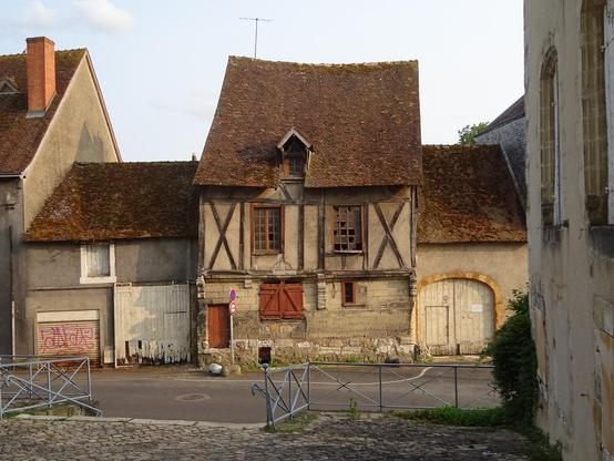 Une maison ancienne avec un toit très pentu vue de face.
Le bas de la bâtisse est en parpaings, une petite porte au palier trop haut, l'étage à colombages avec des fenêtres aux vitres cassées. Elle n'est pas droite mais charmante.