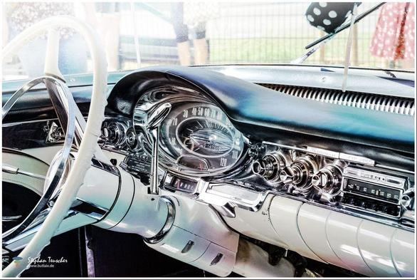 Das Bild zeigt das detailreiche Armaturenbrett eines Oldtimers mit Chrom-Elementen, klassischen Rundinstrumenten, einem großen weißen Lenkrad und nostalgischen Bedienelementen. Im Hintergrund unscharf Personen und eine Wiese.