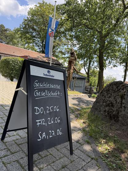 A black A-frame chalkboard sign with white handwriting reads "Geschlossene Gesellschaft" followed by dates: "DO, 25.06.", "FR, 26.06.", and "SA, 27.06.". The sign is placed on a paved path near a building with a flag bearing a blue and white design. Trees and a rock are visible in the background.