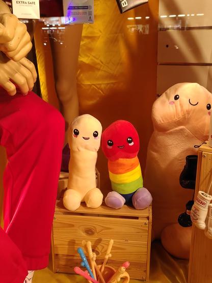 Photo of two cute penis plushies in a shopping window, one in flesh colour of a white person, one in rainbow colours. They have eyes with a pink blush below them and a smiling mouth. Behind them, a larger, flesh-coloured plushie looms in the background. In the front is a cup with penis-shaped pencils.