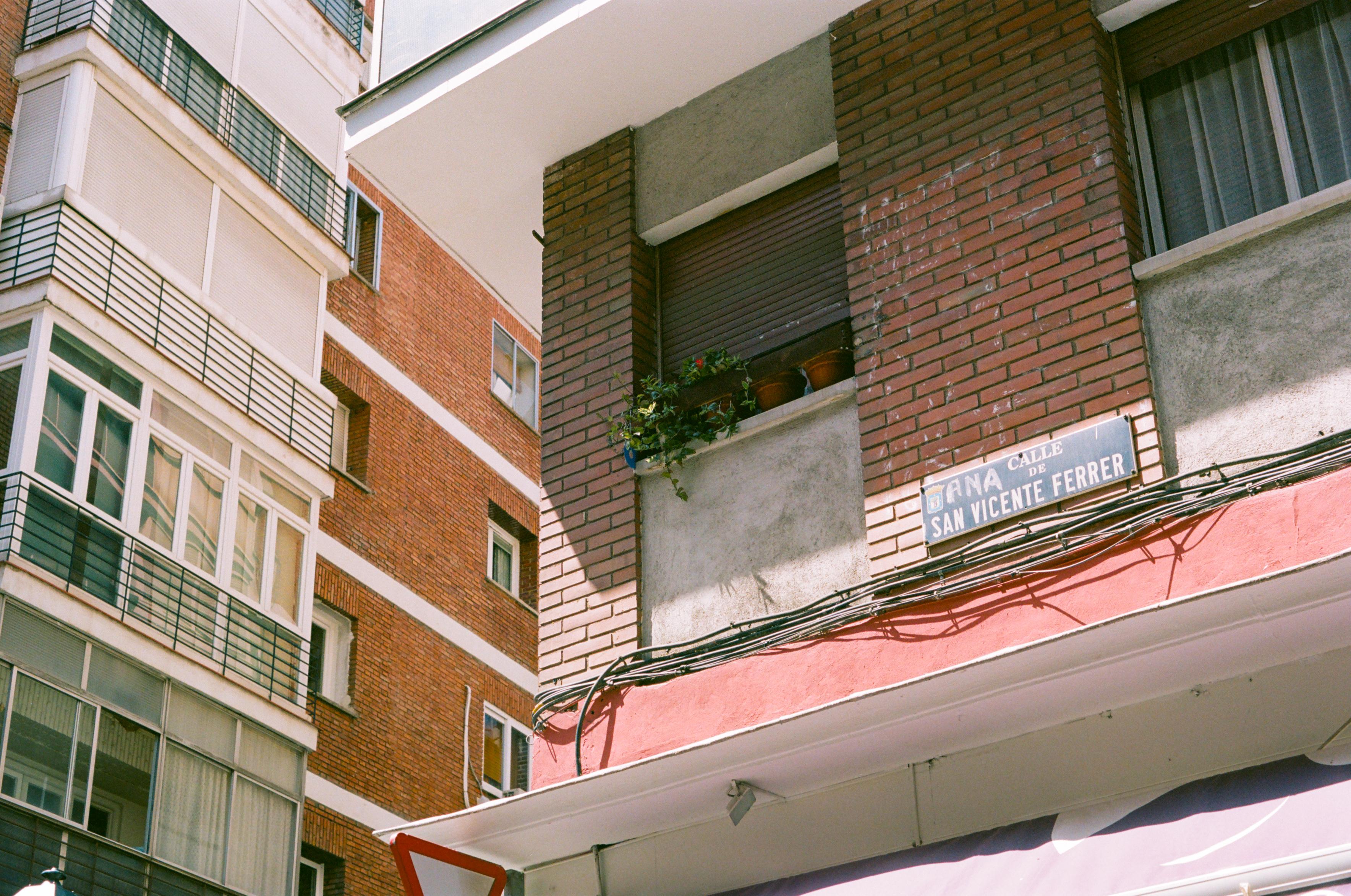 Corner of a red brick building featuring a window in the shade with shutters down and a row of green potted plants. The street plate reads Calle de San Vicente Ferrer. Behind this building, there is another one, and they blend together geometrically.