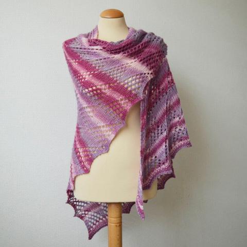 A knit shawl with lace patterning allover in various pinks and lilacs is shown from the front on a mannequin against a white background.