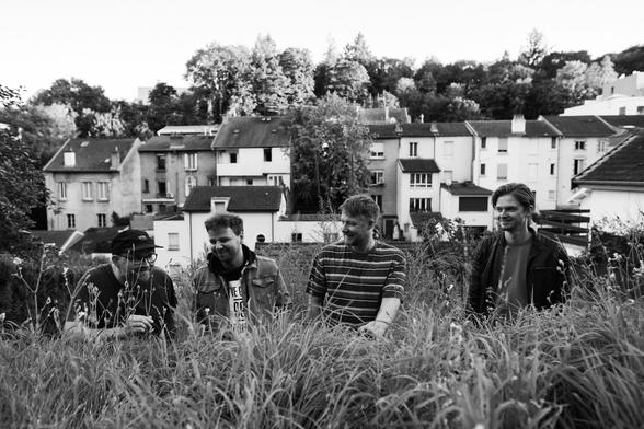 Bandfoto der Paper Planes. schwarz-weiß. Vier junge Männer stehen auf einen mit langem grasbewachsenen Hügel. Etwas hinter dem Gipfel. Die Oberkörper sind halb sichtbar. Im Hintergrund sind Häuser und Bäume!