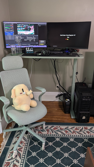 a sage standing desk and office chair. Beside the desk is a large gaming PC with mostly-well managed cables. IN the chair sits a Mastodon plushie. Atop the desk is a keyboard, mouse, Streamdeck keypad, some water, and two large gaming monitors. 
