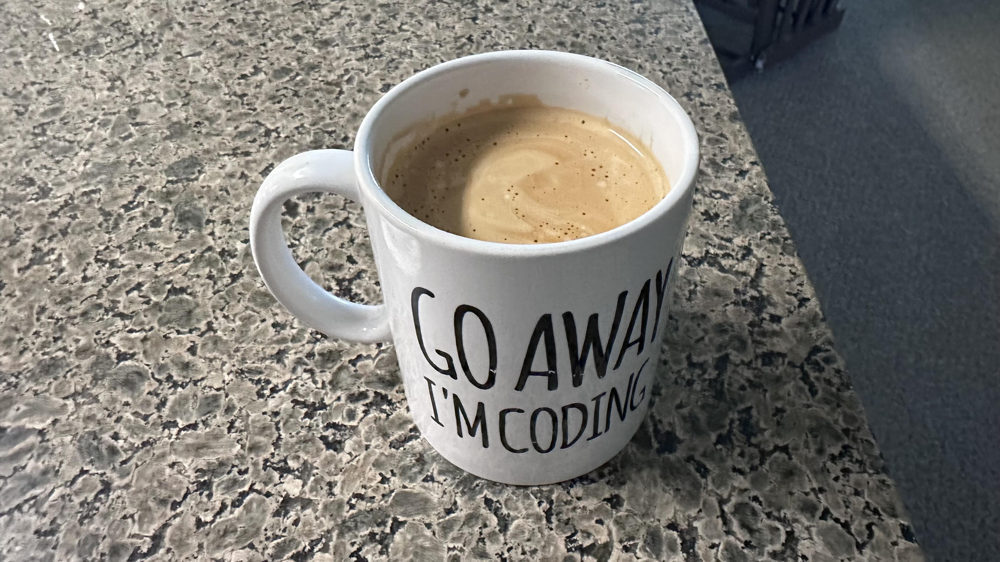 A white mug with the phrase "GO AWAY I'M CODING" filled with coffee, placed on a textured countertop.