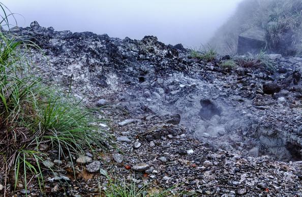 Volcanic gases from a fumarole in a mountainous landscape with grass in the foreground.