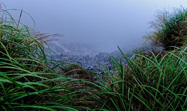 Volcanic gases from a fumarole in a mountainous landscape with grass in the foreground.